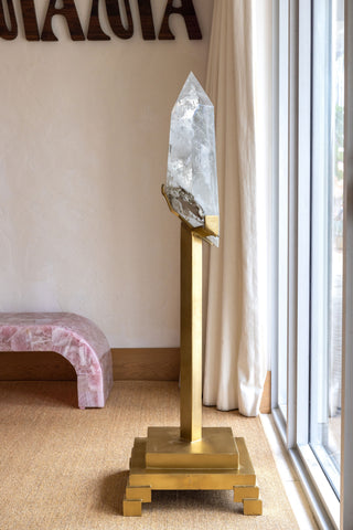 A large clear quartz crystal on a polished gold brass stand, placed in a bright room next to a window with white curtains.