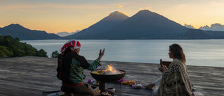 Two people sitting by a fire pit with mountains and lake in the background