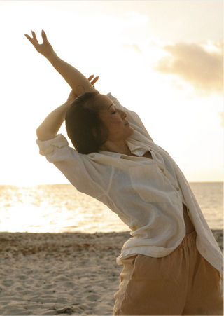 A woman in a white button-up shirt and beige pants stretches her arm over her head on a sandy beach at sunset, with the ocean and warm golden sky in the background.