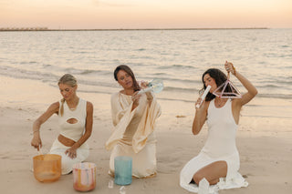 Three women in white performing sacred sound ceremony with singing bowls and crystal pyramid on beach.
