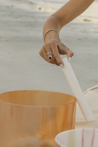 Hand playing citrine aura singing bowl on beach at sunset, sacred jewelry visible on wrist.
