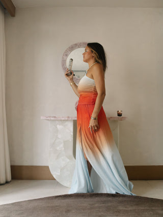 Gabi Torres in flowing orange and white gown standing before rose quartz mirror in atelier.