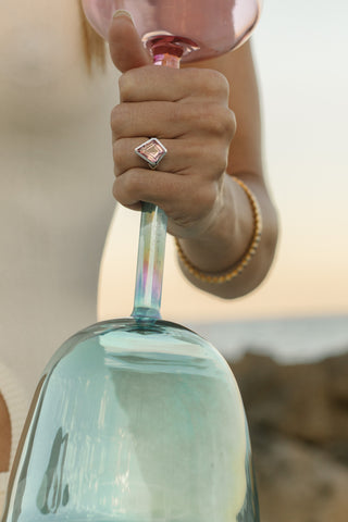 Hand holding aqua and pink aura gold practitioner singing bowl at sunset, beach setting.
