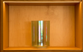 Iridescent aqua aura and lemon gold morph singing bowl displayed on lit wood shelf, front view.
